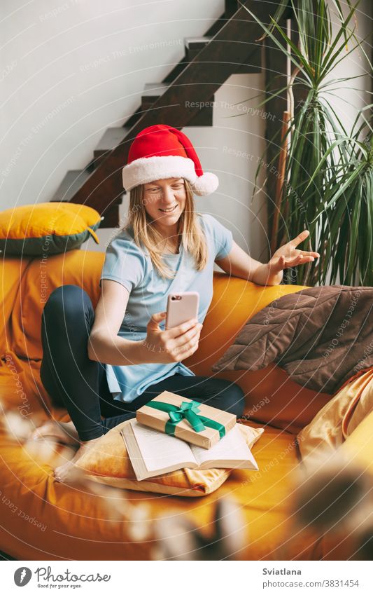 Ein lächelndes Mädchen mit Weihnachtshut sitzt auf dem Sofa und kommuniziert per Videolink mit Freunden. Mädchen gratuliert Freunden und Familie zu den Feiertagen