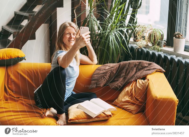 Ein junges Mädchen sitzt auf dem Sofa und telefoniert mit einem Selfie, neben ihr liegt ein offenes Buch heimwärts Frau Funktelefon Raum Telefon Drahtlos Text