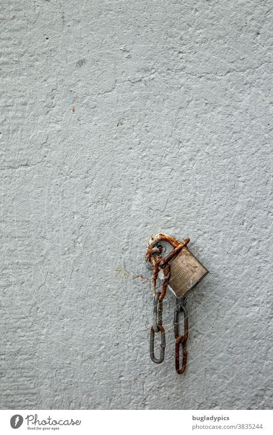 Ein altes Vorhängeschloss/ Schloss hängt zusammen mit einer kurzen Kette an einer Öse an einer weißen Wand. Kettenglieder Rost rostig Eisen Metall Putzwand