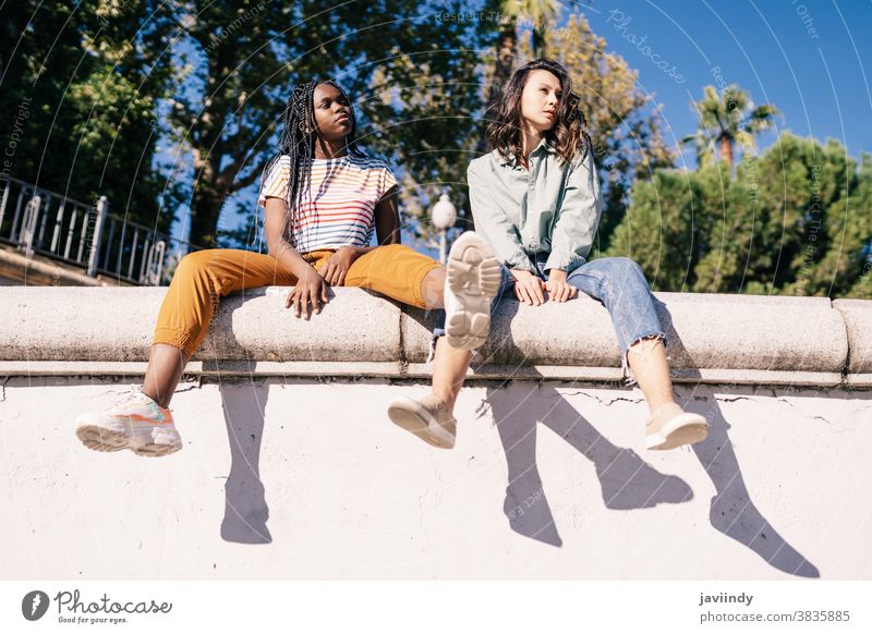 Zwei multiethnische Frauen sitzen auf einer Stadtmauer. ernst Ausdruck Sonnenlicht Freund schwarz Afro-Look Mädchen Schüler zwei Menschen Lifestyle hübsch jung