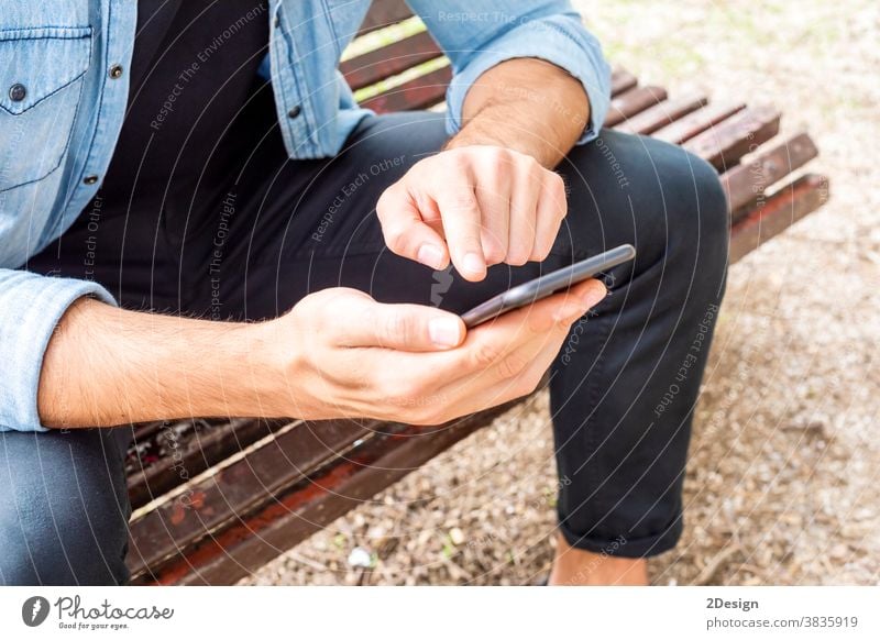 Nahaufnahme eines attraktiven jungen Mannes, der draußen auf einer Bank sitzt und sein Mobiltelefon im Freien benutzt Sitzen Beteiligung Handy Lächeln urban