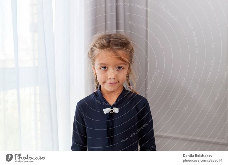 Nahaufnahme eines schönen kaukasischen kleinen Mädchens mit Zöpfen, das mit einem charmanten Lächeln in die Kamera blickt und vor einem hellen Fenster und einer bemalten Wand posiert. Konzept der glücklichen Kindheit