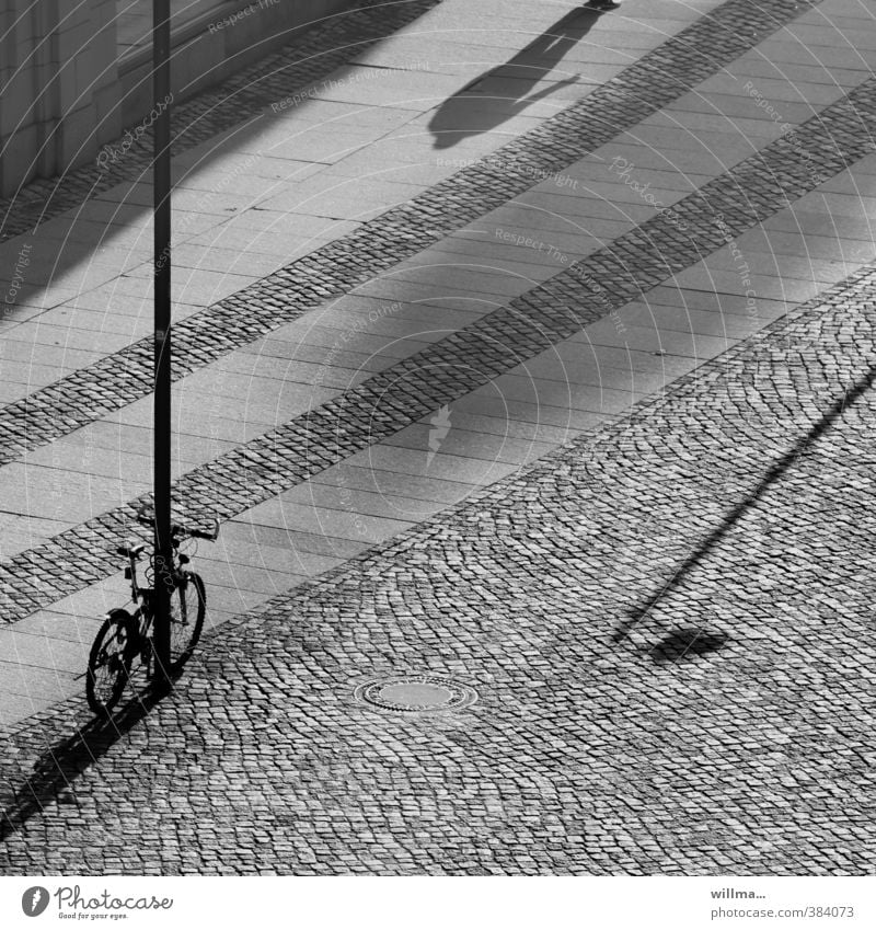 Ein Fahrrad steht, ein Mensch kommt, und der Schatten einer Straßenlaterne drängelt sich ins Bild. Platz Laternenpfahl Schattenspiel sw Pflastersteine