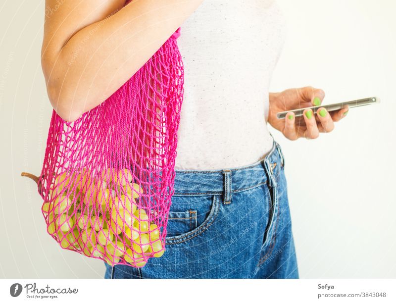 Junge Frau mit wiederverwendbarer Netztasche und Smartphone Tasche ineinander greifen Lebensmittel benutzend Handy online Versand Lebensmittelgeschäft Frucht