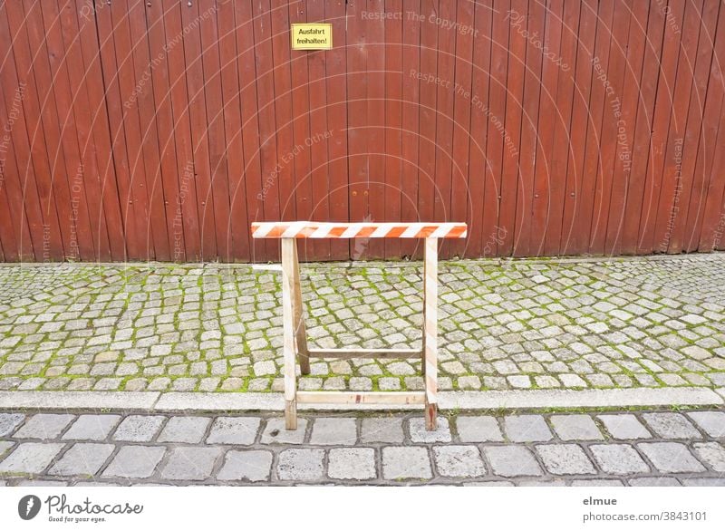 Ein Arbeitsbock aus Holz steht teils auf der Straße, teils auf dem Fußweg vor einem dichten, rot-braunen Holzlattenzaun mit einem gelben Schild "Ausfahrt freihalten!"