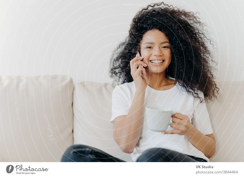 Horizontale Aufnahme einer gut aussehenden Frau, die ein Ferngespräch per Mobiltelefon führt, während des Gesprächs informelle Fragen löst, im Wohnzimmer auf dem Sofa sitzt, Tee trinkt, ein breites Lächeln hat, weiße Zähne zeigt