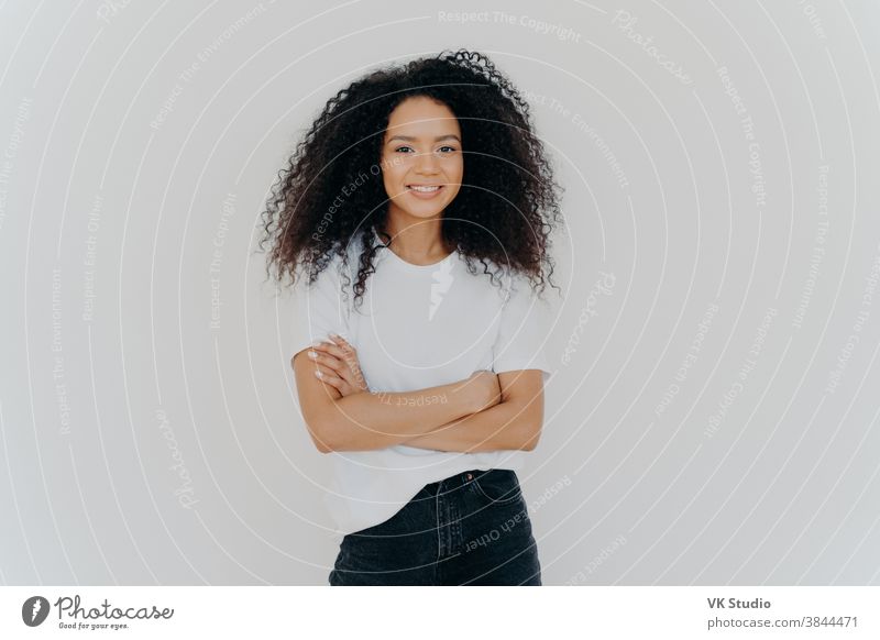 Studioaufnahme einer unbekümmerten jungen, schönen Frau mit Afro-Frisur, die die Arme verschränkt hält, fröhlich lächelt, lässiges T-Shirt und Jeans trägt, isoliert auf weißem Hintergrund. Menschen, Ethnizität, Gesichtsausdrücke