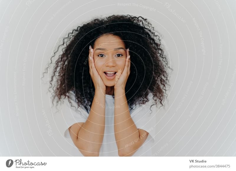 Foto einer überraschten fröhlichen Frau mit Afro-Haarschnitt, hält beide Hände auf den Wangen, hat natürliche Schönheit, öffnet den Mund, kann nicht an aufregende Nachrichten glauben, trägt T-Shirt, posiert vor weißem Hintergrund.