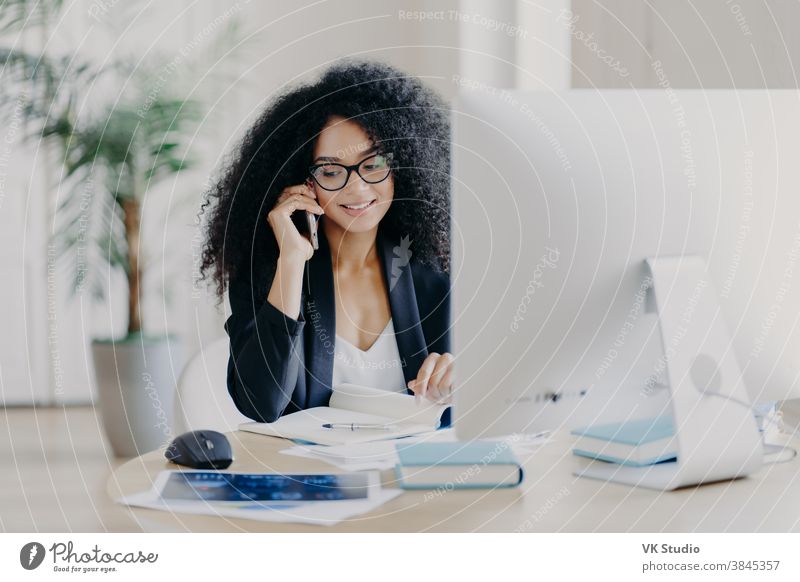 Foto einer zufriedenen Afroamerikanerin, die per Smartphone telefoniert, Notizen im Notizblock macht, am Arbeitsprozess beteiligt ist, am Schreibtisch mit Computer sitzt, eine Brille trägt, formelle Kleidung trägt, telefoniert