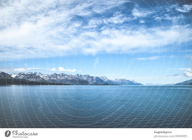 Ein Fjord in Norwegen. Im Vordergrund das Meer und im Hintergrund schneebedeckte Berggipfel. Den blauen Himmel zieren Schleierwolken. Küste Berge u. Gebirge