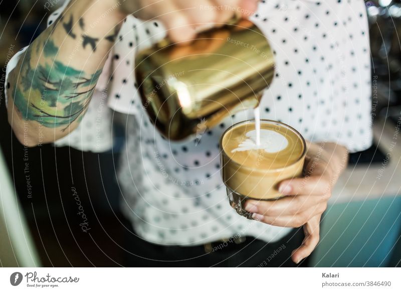 Tätowierte Barista gießt mit Milchschaum ein Herz in ein Glas Milchkaffee mit Latte Art in der Hand gießen milchschaum café au lait Espresso Latte Macchiato
