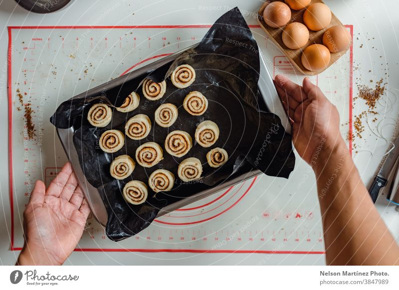 Die Hand eines Mannes hält ein Tablett mit Zimtbrötchen, die bereit sind, in den Ofen geschoben zu werden. im Innenbereich Essen zubereiten Teigwaren Mehl