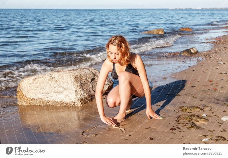 junges Mädchen im Badeanzug zeichnet mit dem Finger auf den Sand am Meeresufer zeichnen Schönheit Glück schön Zeichnung sonnig Frau Wasser Küste Sonne Feiertag