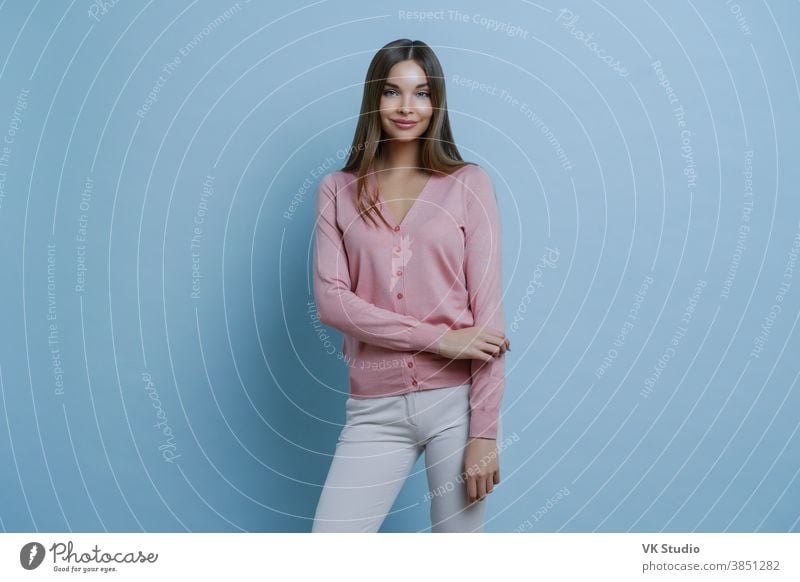 Modische brünette Kaukasierin in rosa Pullover und weißer Hose, geht zur Arbeit, schminkt sich, lächelt angenehm, isoliert auf blauem Hintergrund. Menschen, Stil, Lifestyle-Konzept
