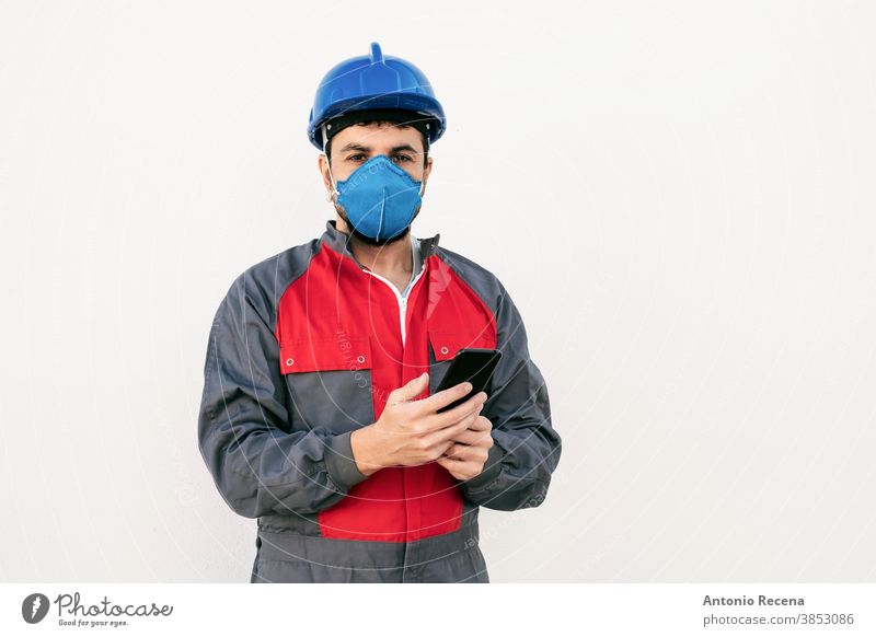 Bärtiger Arbeiter Mann mit Gesichtsmaske sieht Smartphone Telefon covid-19 Mundschutz Uniform Sicherheit Pandemie Virus weißer Hintergrund Wand Coronavirus