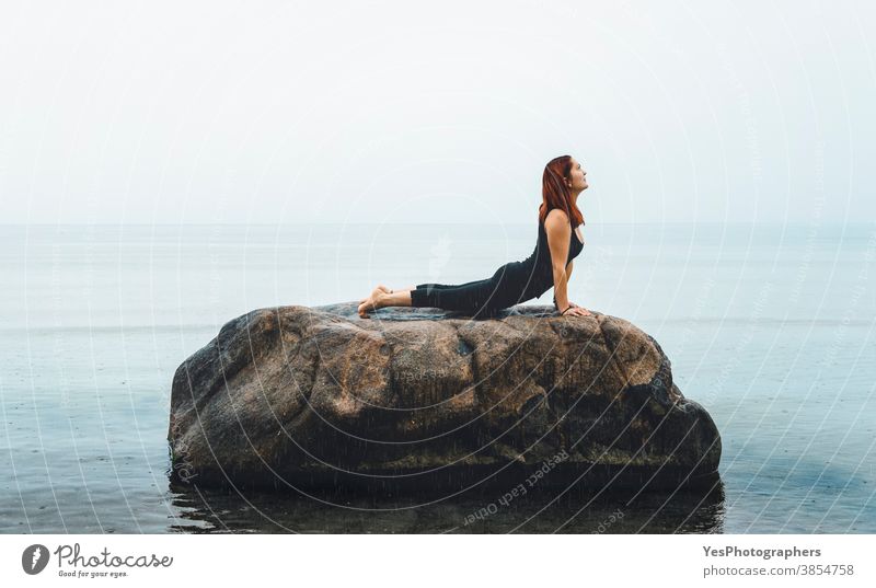 Junge Frau, die Yoga praktiziert, auf See, im Regen. Rothaariges Mädchen meditiert auf einem Felsen in der Ostsee, Deutschland 20s Iyengar-Yoga Erwachsener