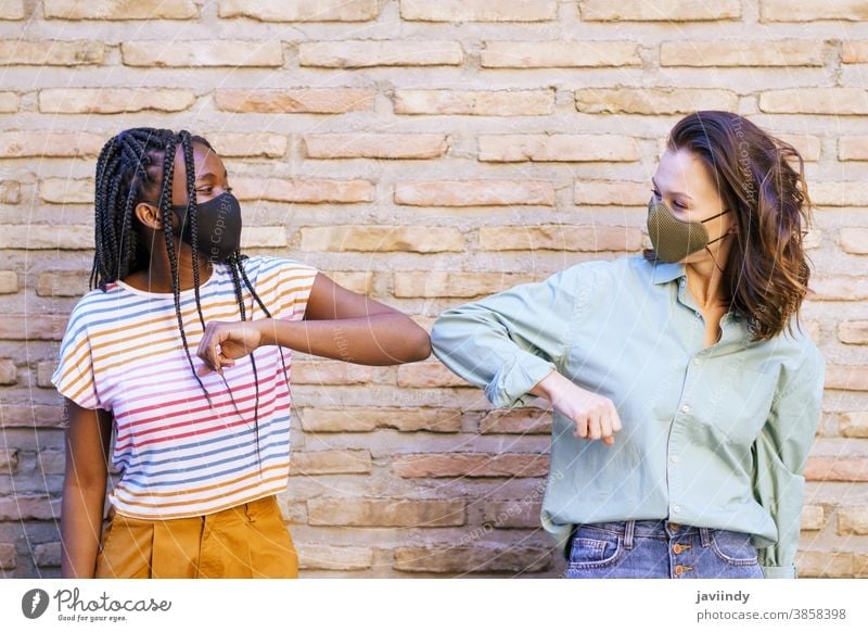 Multiethnische junge Frauen mit Masken, die einander mit den Ellbogen grüßen Begrüßung Menschen sozial Distanzierung Mundschutz Pandemie Virus Coronavirus