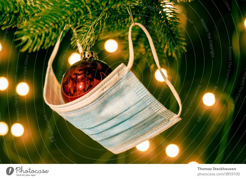 Rote Christbaumkugel und eine Mundschutzmaske auf einem Ast vom Weihnachtsbaum, im Hintergrund Lichter. Symbol für die Auswirkungen des Covid-19-Virus in der Weihnachtszeit 2020 und 2021