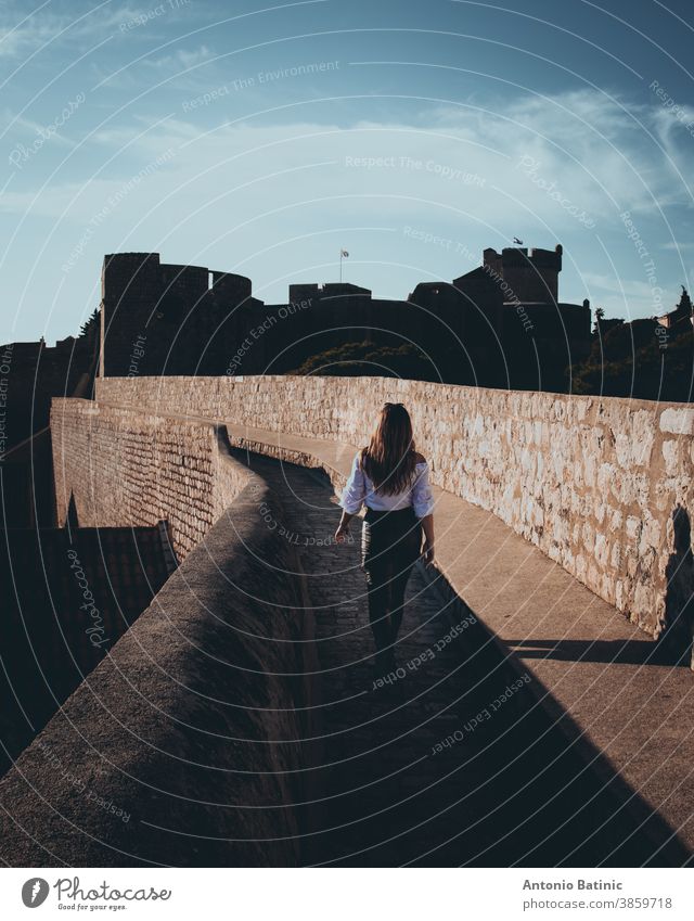 Vertikalaufnahme und Rückansicht eines attraktiven Mädchens, das entlang der Stadtmauer der Stadt Dubrovnik läuft. Dunkler stimmungsvoller Schnitt mit schwarzen und orangefarbenen Highlights