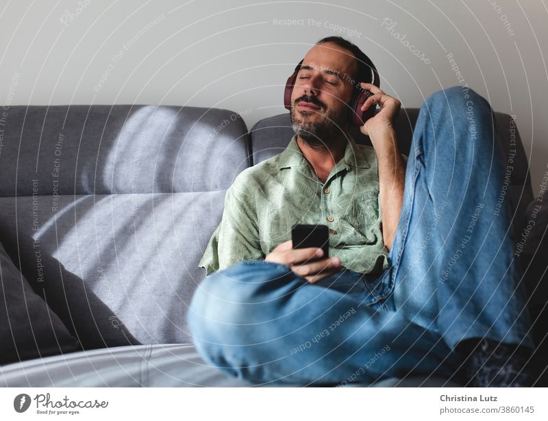 Mann sitzt auf dem Sofa im Wohnzimmer und hört Musik mit Kopfhörern zuhören Sitzen sich[Akk] entspannen Telefon heimwärts Menschen Mobile Person Mitteilung
