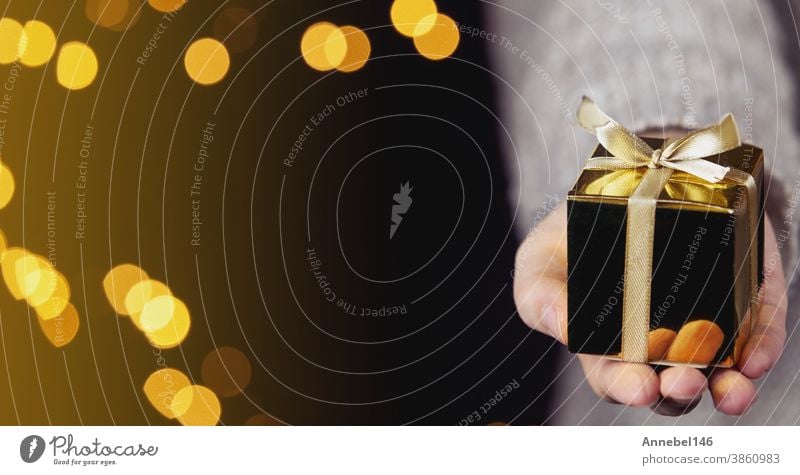 Hand hält eine goldene Geschenkverpackung mit Goldband, Bokeh-Glitzer-Hintergrund, Weihnachts- oder Geburtstagsgeschenkkonzept mit Platz für Text Feiertag