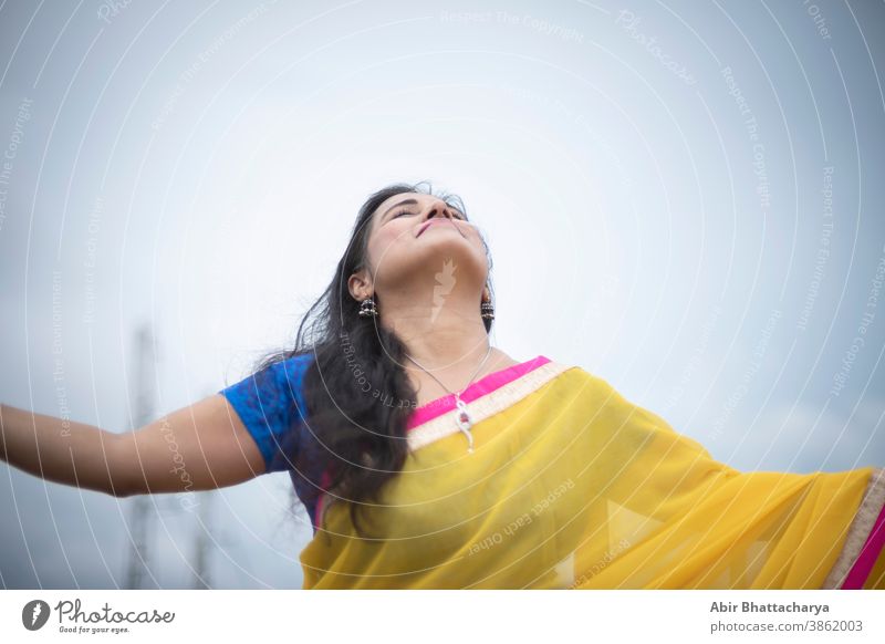 Eine junge und schöne indisch-bengalische brünette Frau in traditionellem gelben Sari und blauer Bluse steht, während sie ihre Arme auf dem Dach unter blauem Himmel mit Wolken ausbreitet. Indischer Lebensstil