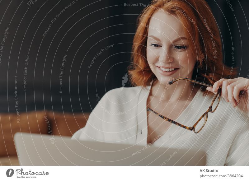 Schöne rothaarige Frau konzentriert auf Laptop-Display, lächelt positiv, hält optische Brille, posiert in Innenräumen, nutzt moderne Technologien für Online-Kommunikation, schaut Webinar, liest Nachrichten