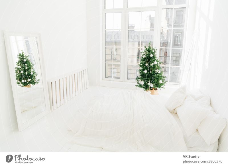 Komfort-, Haus- und Heiligabend-Konzept. Schlafzimmer mit weißem Softbett, Spiegel, großem Fenster für Lichteinfall, dekoriertem Neujahrsbaum und Leiter. Feiertags-Dekoration.