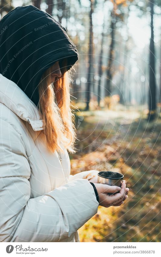 Frau mit Kapuze, die während einer Herbstreise eine Tasse mit heißem Getränk aus der Thermoskanne an einem kalten Herbsttag in der Hand hält. Aktives Mädchen wandert in einem Wald und verbringt aktiv Zeit