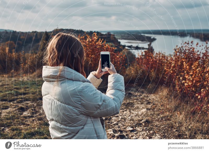Rückansicht einer jungen Frau, die während einer Reise an einem sonnigen Herbsttag mit einem Smartphone Fotos von der Landschaft macht Junge Frau nehmen unter