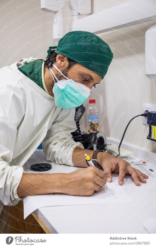 Arzt macht sich Notizen im medizinischen Bericht zur Kenntnis nehmen Mann schreiben Schriftstück Sanitäter Mundschutz Klinik männlich Krankenhaus Tisch Raum