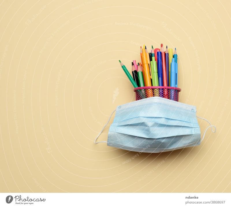medizinische Einwegmaske und mehrfarbige Stifte und Bleistifte in einem Metalleimer auf beigem Hintergrund Mundschutz Pandemie Schreibstift Prävention schützend