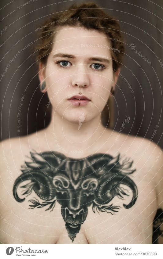 Portrait einer jungen Frau, die eine großflächige Tätowierung eines Ochsenkopfes auf den Dekolletè hat dunkelblond Schmuck Piercing Ohrring Dekolleté Brust