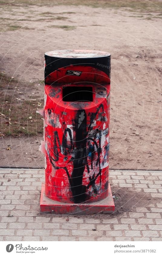 Ein wunderschöner roter eiserner Abfallbehälter mit vorderem Einwurf.  Etwas verschmiert mit schwarzer Farbe, steht einsam und verlassen auf dem Bürgersteig. Das Wort NO kann man noch erkennen.