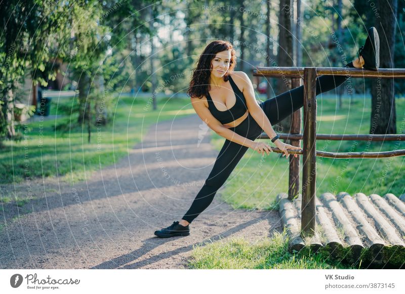 Flexible junge gelockte Frau trägt schwarze Top-Leggings und Turnschuhe, streckt die Beine, treibt an Sommertagen Sport an der frischen Luft, posiert vor grünen Bäumen. Menschen, Flexibilität und Übungskonzept