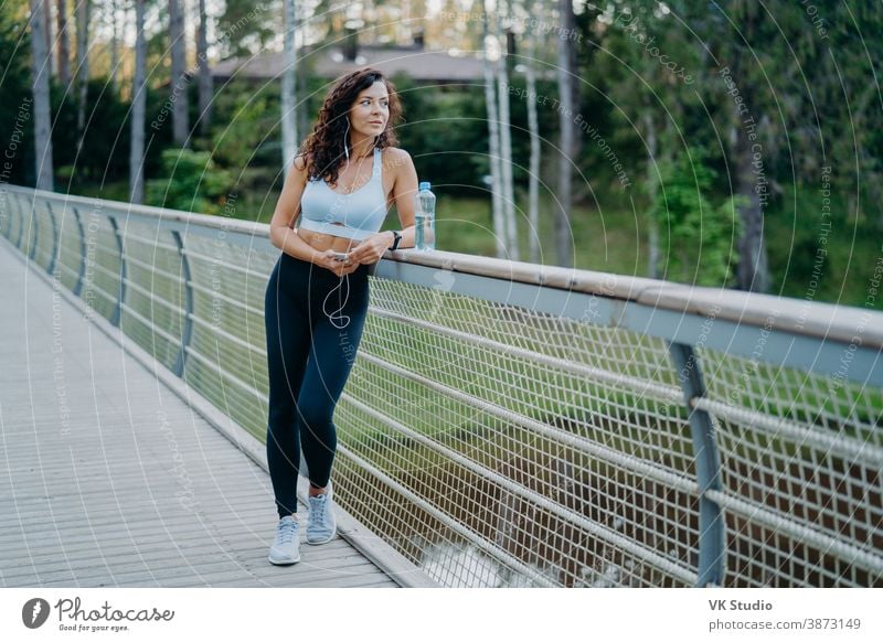 Seriöse nachdenkliche Frau mit fleckigem Körper, bekleidet mit kurz geschnittenem Top und Leggings, posiert an der Brücke, schaut nachdenklich weg, hört Musik in Kopfhörern. Menschen, gesunde Lebensweise und Fitnesskonzept