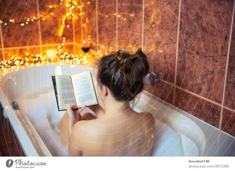 Junge schöne Frau liest ein Buch und trinkt Rotwein in der Badewanne mit Sprudelschaum, dekoriert mit bunten Lichtern, Entspannungs- und Spa-Konzept Wein jung