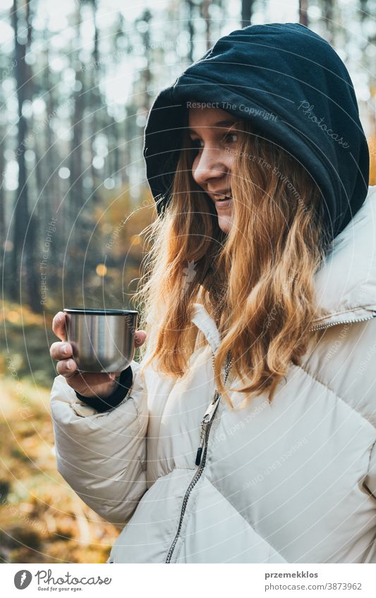 Frau mit Kapuze, die während einer Herbstreise Pause macht und am kalten Herbsttag eine Tasse mit heißem Getränk aus der Thermoskanne hält aktiv Aktivität