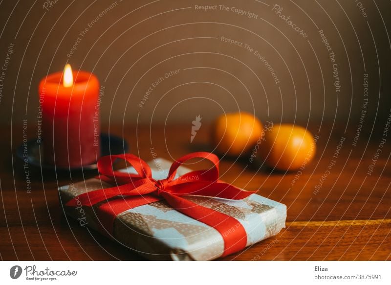 Ein Weihnachtsgeschenk liegt auf dem Tisch, daneben zwei Mandarinen und eine brennende rote Kerze. Weihnachten Advent schenken Bescherung Nikolaus Kerzenschein