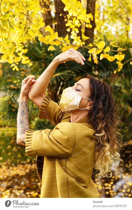 Frau in gelbem Kleid und Maske in einem Park retuschieren Behaarung Mundschutz Garten Gelblich Blätter Lifestyle reif Porträt eine Person Baum Mantel
