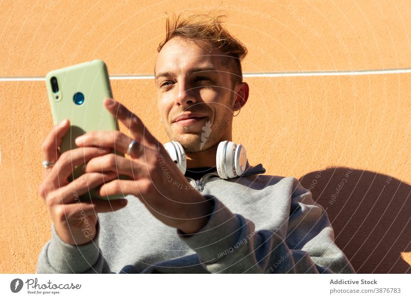 Junger Hipster-Mann beim Surfen auf dem Smartphone benutzend Wand urban Browsen positiv Telefon online jung Typ lässig Kopfhörer Mobile sonnig Schatten