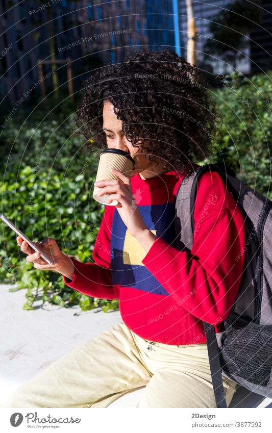 Hübsche junge Afro-Frau sitzt auf einer Bank im Freien, benutzt ihr Mobiltelefon und trinkt eine Kaffeetasse zum Mitnehmen Sitzen Mobile Smartphone Mitnahme