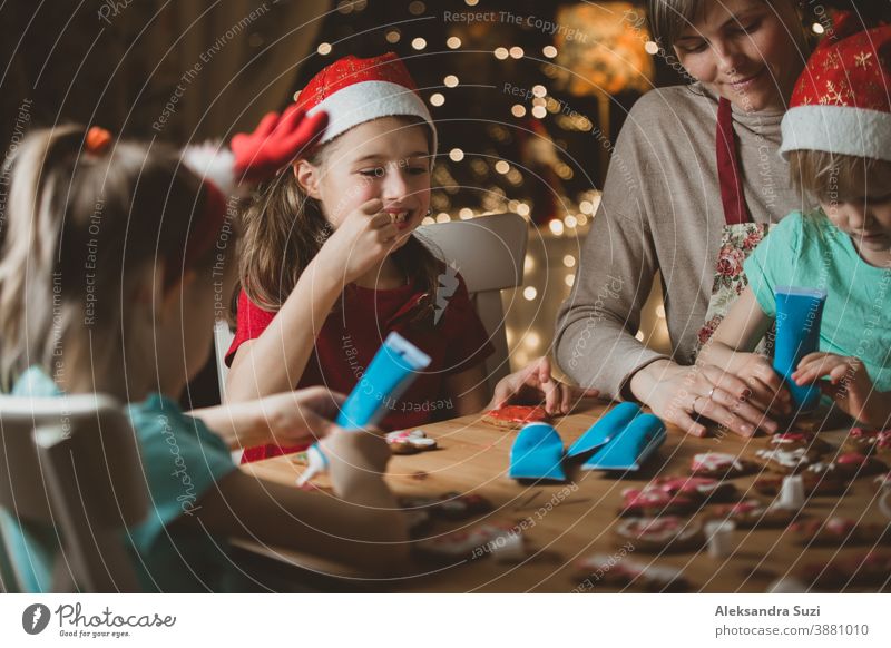 Mutter und kleine Kinder mit roten Hüten, die Lebkuchen backen und mit Glasur dekorieren. Wunderschönes Wohnzimmer mit Lichtern und Weihnachtsbaum. Fröhliche Familie, die gemeinsam Feiertage feiert.