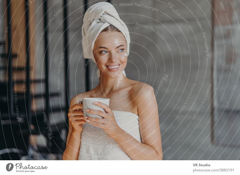 Attraktive junge Frau trägt nach dem Duschen Feuchtigkeitscreme auf das Gesicht auf, lächelt breit, in Handtücher gehüllt, trinkt Kaffee, posiert drinnen, denkt an etwas Angenehmes. Konzept der Schönheitspflege