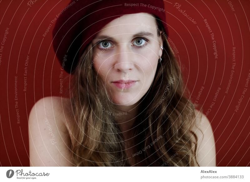 Portrait einer jungen Frau mit Hut vor einer roten Wand schlank schön brünett lange Haare Gesicht schlau emotional sehen schauen Blick direkt natürlich