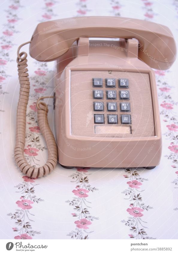 Ein altes Telefon in rosa steht auf einem Tisch mit Blumenmuster. Vintage, fortschritt, Technik, kommunikation vintage tasten Zahlen tapete blumenmuster