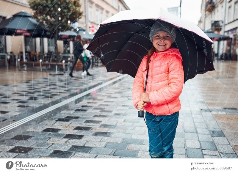 Kleines Mädchen mit großem Regenschirm in der Hand geht an einem verregneten, düsteren Herbsttag durch die Innenstadt im Freien wenig saisonbedingt fallen