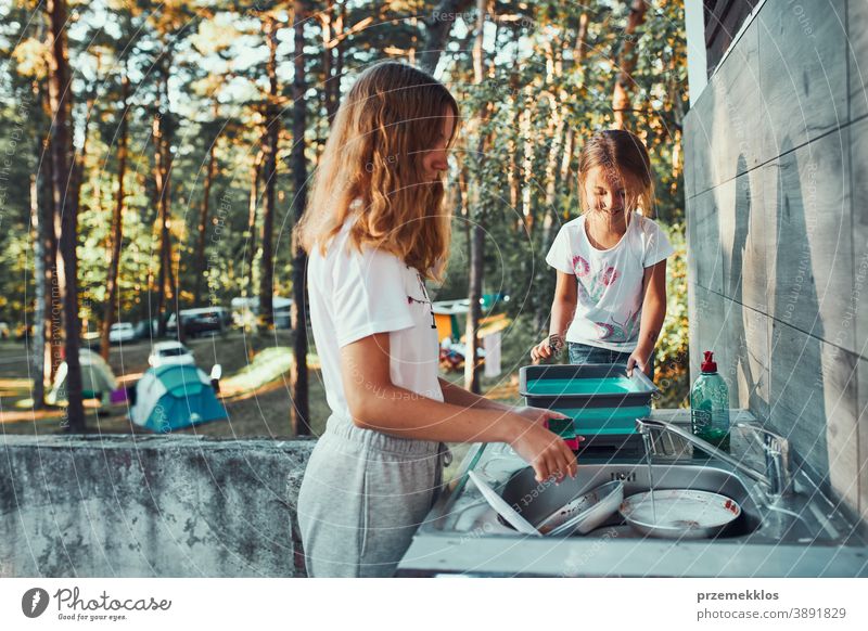 Teenager-Mädchen spült während der Ferien auf dem Campingplatz mit Hilfe ihrer jüngeren Schwester in der Außenküche Töpfe und Teller ab arbeiten Zusammensein