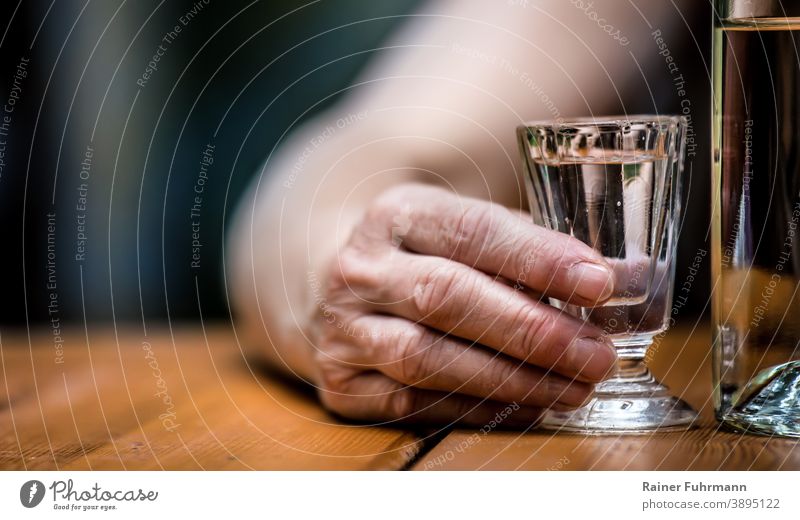 Auf einem Tisch stehen eine Flasche Alkohol und ein Schnapsglas. Die Hand einer Person hält das Schnapsglas fest. anonym Sucht süchtig betrunken Alkoholsucht