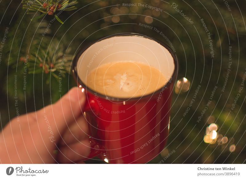 Adventskaffee. Hand hält roten Becher mit Cappuccino. Nahaufnahme mit Tannengrün und einigen glänzenden unscharfen Lichtern im Hintergrund Kaffeebecher